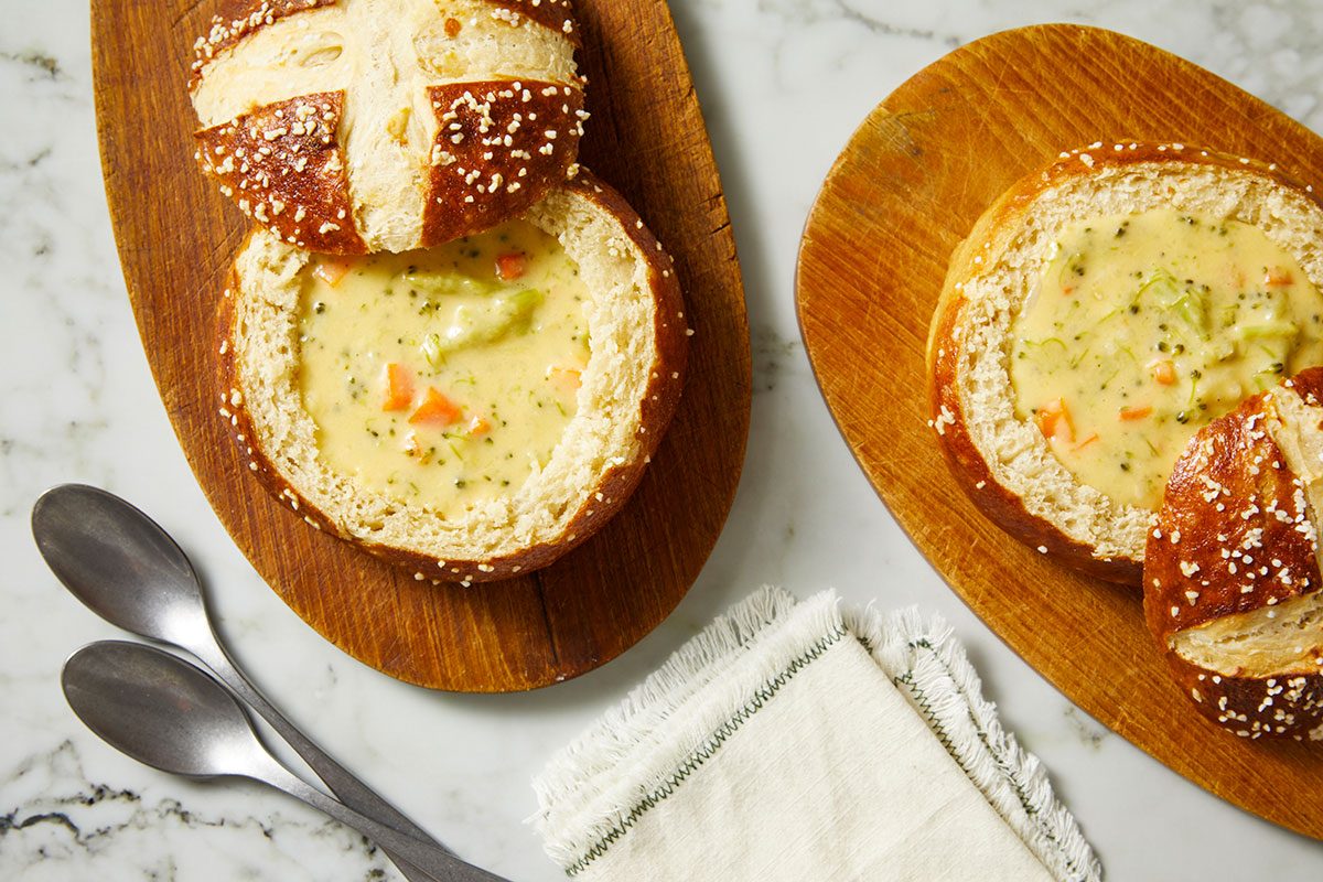 Overhead shot of two bread bowls filled with creamy vegetable soup on wooden boards, with two metal spoons and a folded white napkin nearby on a marble surface.