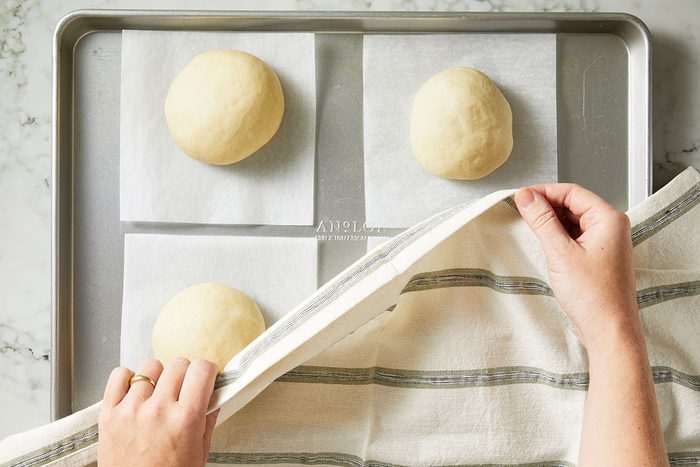 Overhead shot of three dough balls on parchment squares resting on a baking sheet; A person’s hands, one wearing a gold ring, lift a striped kitchen towel partially covering the dough, set on a light marble surface.