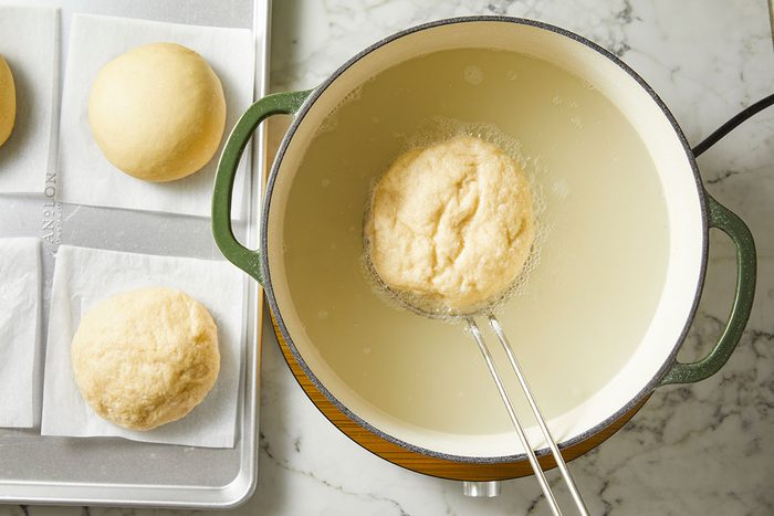 Overhead shot of a round dough ball being lifted from a pot of hot oil with tongs, while three more dough balls rest on parchment paper on a baking sheet nearby; The scene is set on a marble countertop.