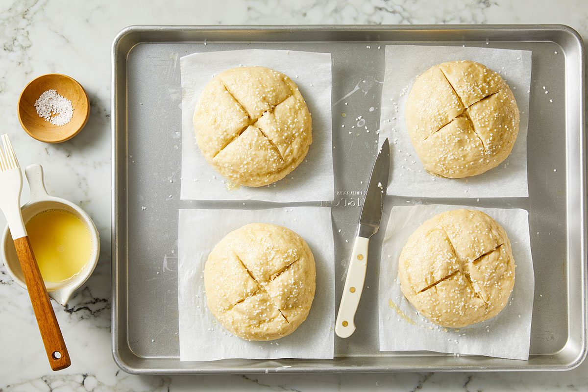 Overhead shot of a baking sheet with four round, unbaked dough loaves scored with an X and placed on parchment squares; Nearby are a knife, a pastry brush, a bowl of melted butter, and a small wooden bowl of salt.