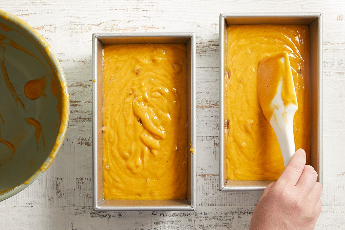 A hand spreads yellow batter in a rectangular loaf pan with a spatula, next to another pan already filled with batter. A mixing bowl with leftover batter sits nearby on a white wooden surface.