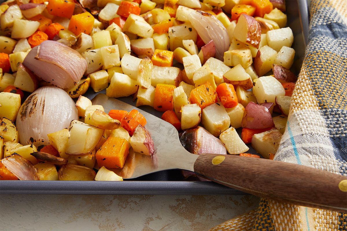 A baking tray filled with roasted root vegetables, including diced potatoes, carrots, parsnips, and chunks of red onion, with a metal spatula and a plaid cloth nearby.