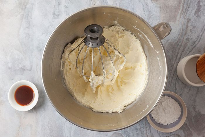 Overhead shot of a mixing bowl filled with whipped butter and sugar, surrounded by a whisk attachment, a small bowl of vanilla extract, and a bowl of powdered sugar on a light countertop