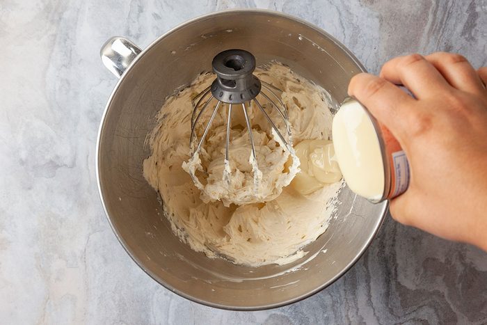 Overhead shot of a hand pouring sweetened condensed milk from a can into a mixing bowl of whipped cream, with a stand mixer fitted with a whisk attachment.