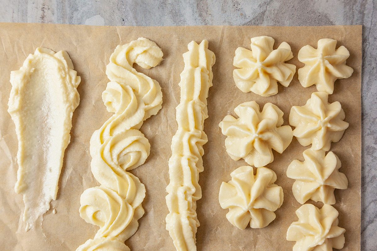 Overhead shot of various Russian buttercream piping styles on parchment paper, featuring smooth swipes, elegant swirls, ridged lines, and flower-shaped dollops that showcase the creamy, pale buttercream against a warm-toned background.