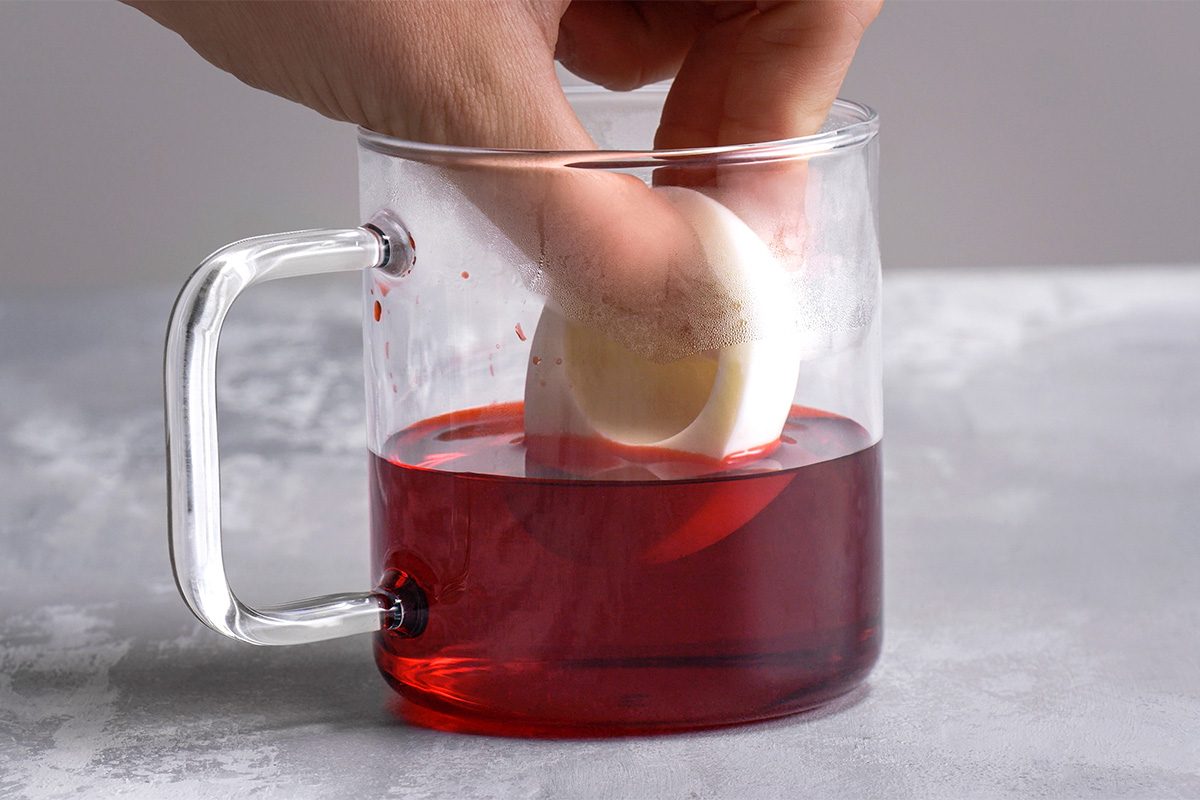 A hand dipping a white egg into a clear glass mug filled with red liquid, likely for egg dyeing, on a light gray surface.