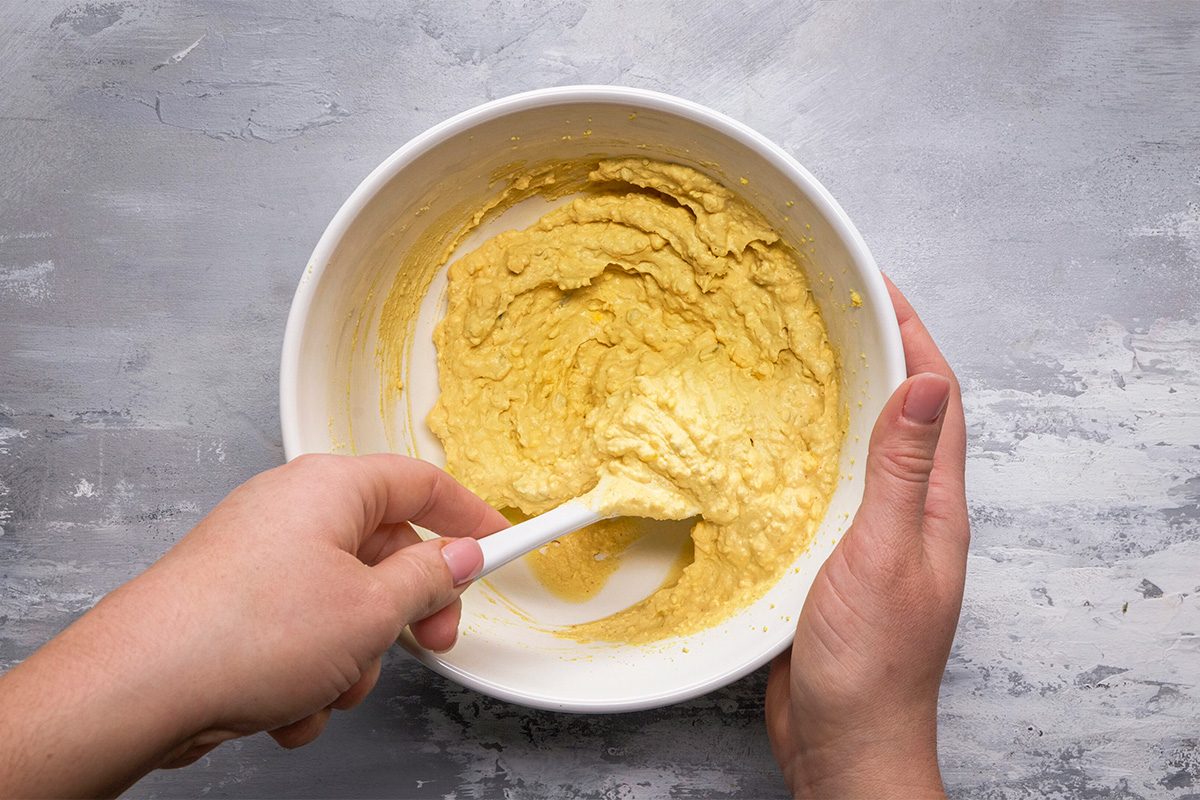A person holding a white bowl and stirring yellow batter with a white spatula on a gray textured surface.