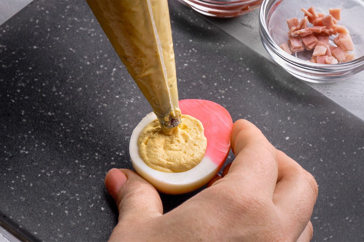 A hand holds a halved pickled egg with a pink edge while a piping bag fills the center with a yellow deviled egg mixture; diced ham and ingredients are in glass bowls nearby.