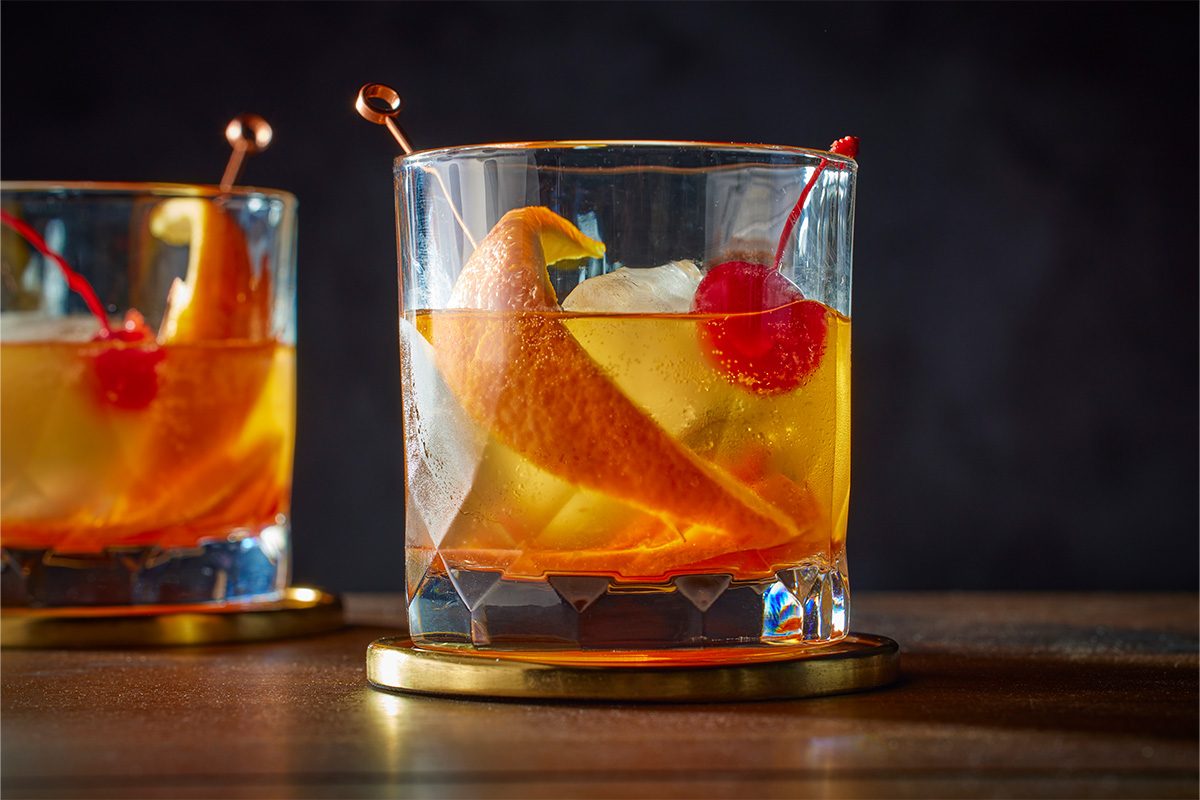 An eye level shot of a Scotch Old-Fashioned, a glass with ice, orange peel, and a maraschino cherry on a cocktail pick, resting on a gold-rimmed coaster, with another drink visible behind;