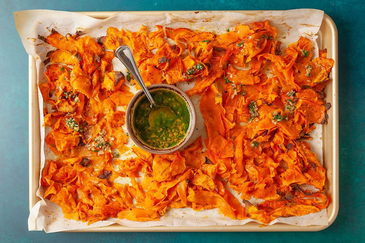 Overhead shot of a parchment-lined sheet pan holding roasted, thinly sliced sweet potatoes, crisped around the edges, with a small bowl of green herb sauce and a spoon placed in the center;