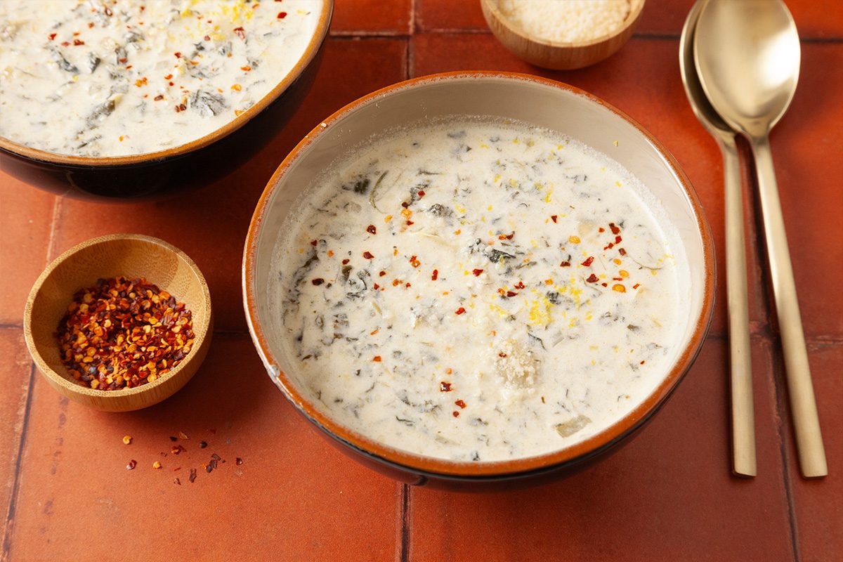 Overhead shot of a bowl of Spinach Artichoke Dip Soup topped with herbs and red pepper flakes, placed near small bowls of pepper flakes, grated cheese, and two spoons;
