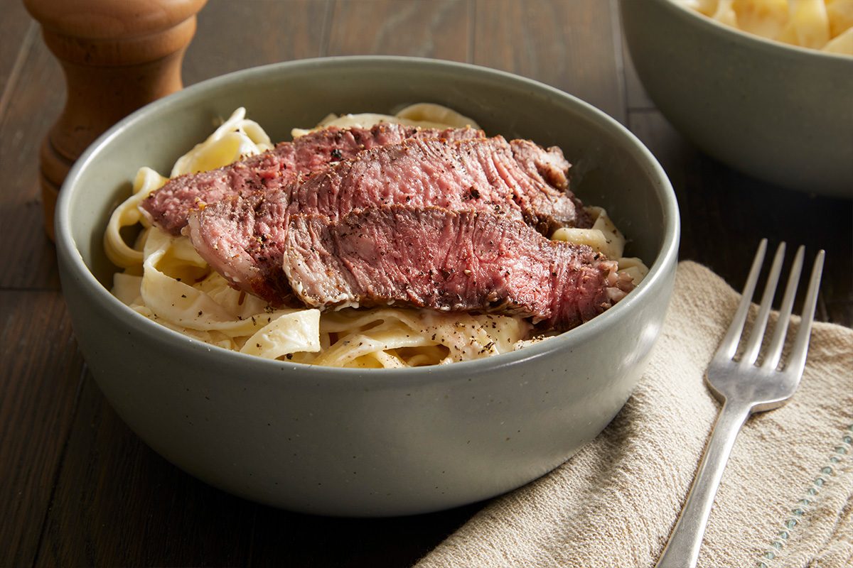 Overhead shot of sliced steak arranged neatly over Alfredo pasta in a gray bowl with a fork on the side.