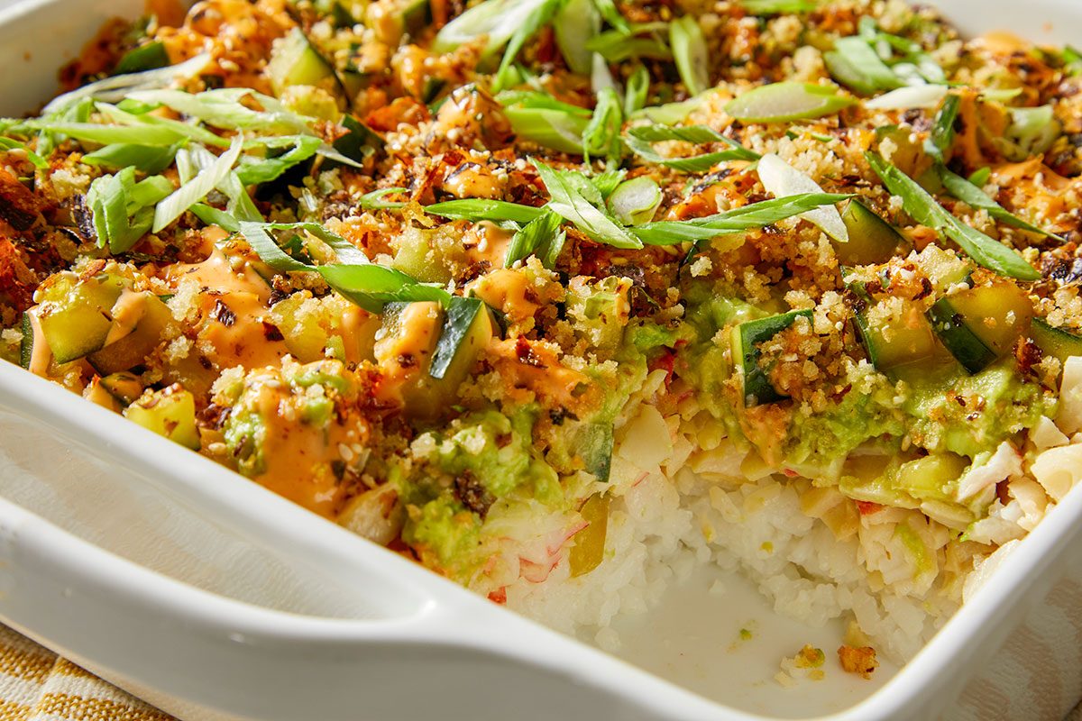 Closeup shot of Sushi Bake; a scooped-out section reveals layers of fluffy white rice, imitation crab, creamy sauce, and diced cucumber; the top is covered with toasted breadcrumbs, spicy mayo, and sliced green onions; served in a white baking dish;