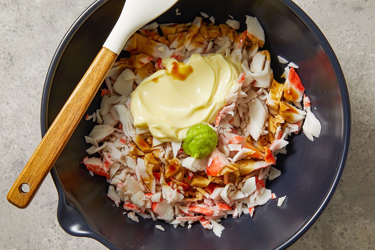 Overhead shot of a black mixing bowl containing chopped imitation crab; a dollop of mayonnaise; a small scoop of green wasabi; and a splash of soy sauce; a white spatula rests on the edge of the bowl;