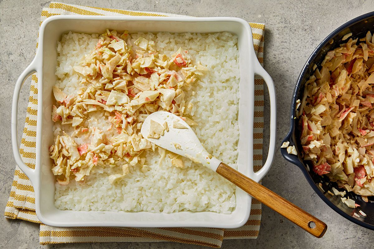 Overhead shot of a white baking dish filled with a layer of cooked rice; partially topped with a creamy imitation crab and vegetable mixture; a spatula rests in the dish; a pan with more of the mixture sits nearby on a striped towel;