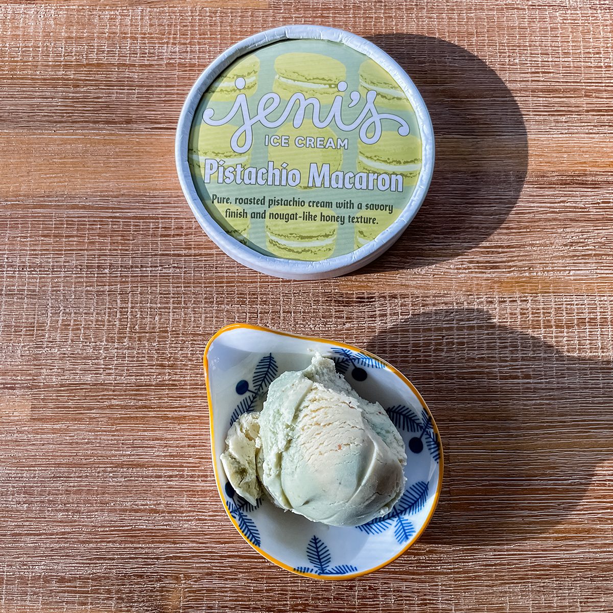 A small bowl with two scoops of pale green pistachio macaron ice cream sits beside a container labeled "Jeni's Ice Cream Pistachio Macaron" on a wooden surface.