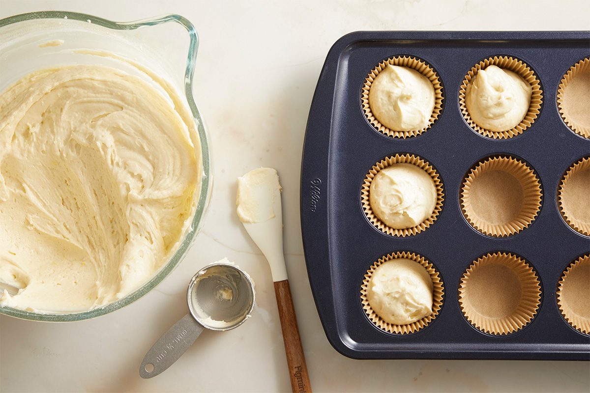 A bowl of cupcake batter, a spatula, a metal scoop, and a muffin tin lined with paper baking cups, some filled with batter, are arranged on a light countertop.