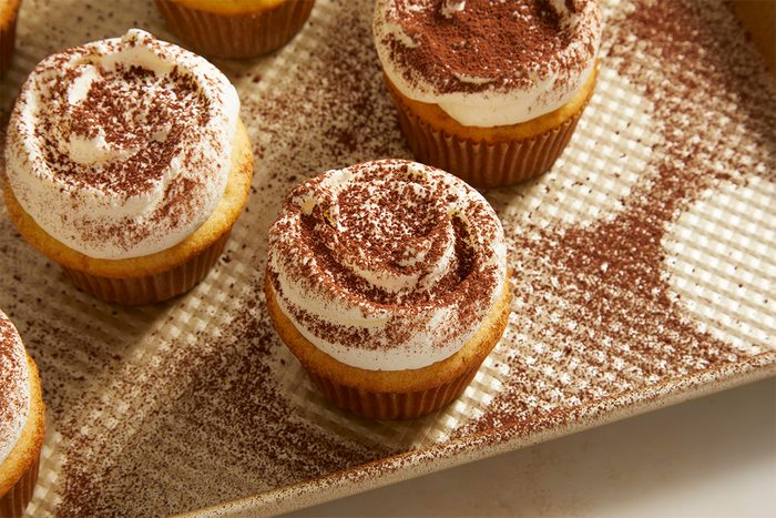 Cupcakes topped with swirls of white frosting and dusted with cocoa powder, arranged on a textured baking tray with cocoa powder scattered around them.