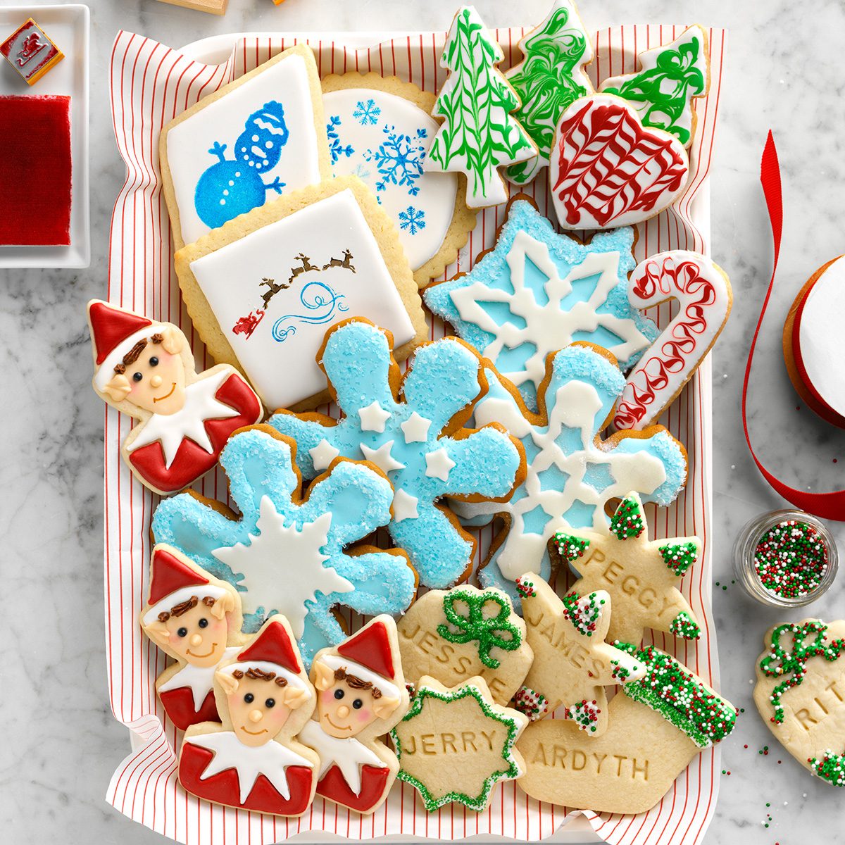 A tray of decorated holiday cookies, including snowflakes, elves, trees, candy canes, and gift tags with names. The cookies are colorful with red, green, blue, and white icing. A red ribbon and icing bowl are nearby.
