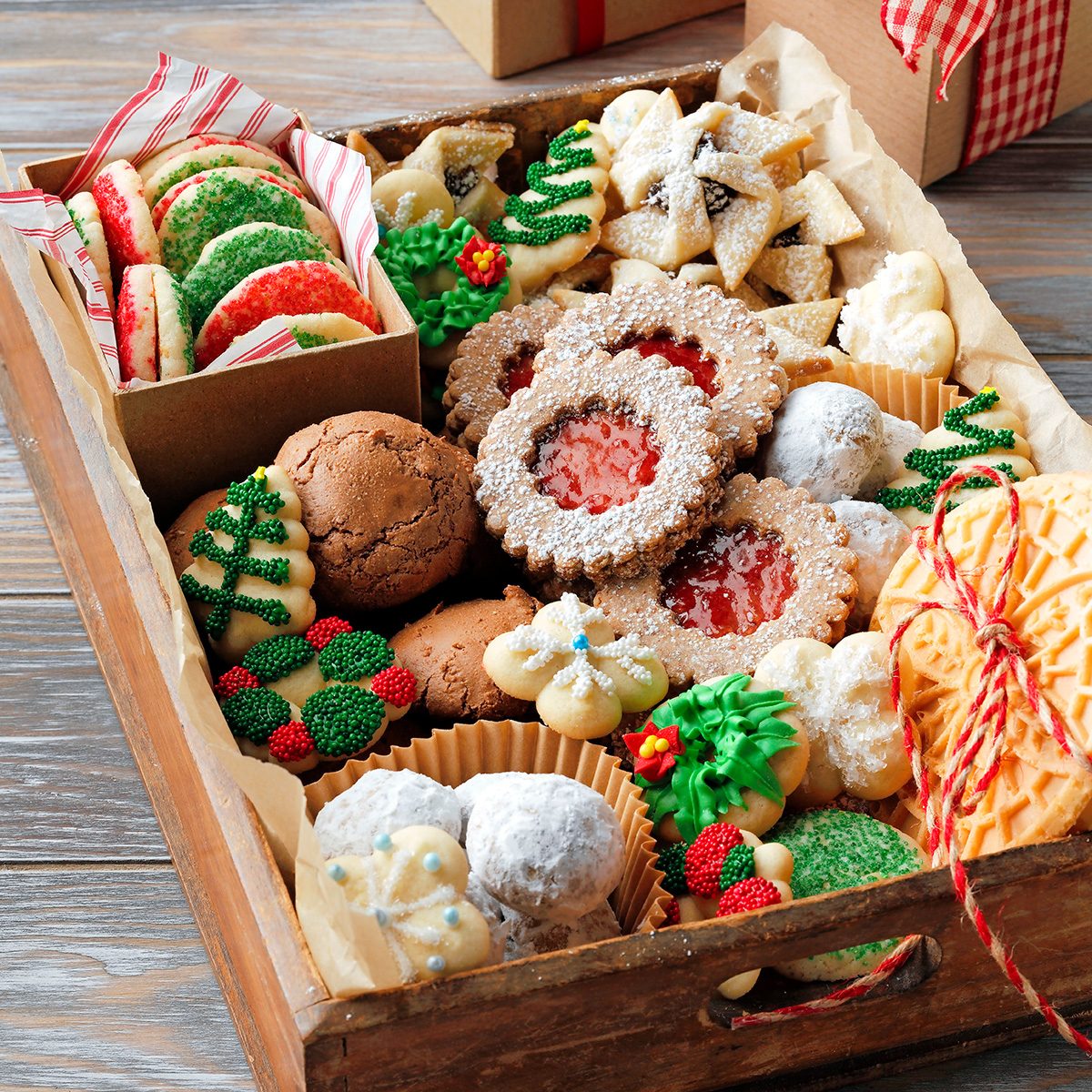 A wooden tray filled with assorted colorful holiday cookies, including decorated sugar cookies, jam-filled linzers, biscotti, and round powdered sugar cookies, arranged neatly with festive decorations.