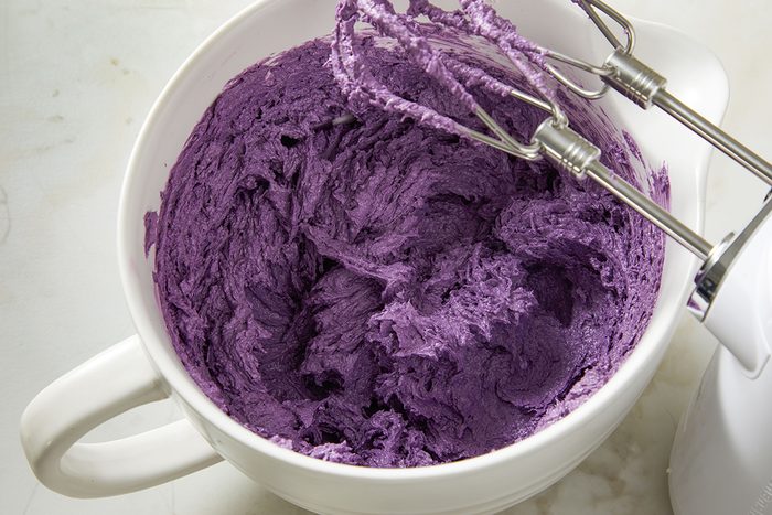 A white mixing bowl filled with vibrant purple batter or frosting, being mixed with an electric hand mixer. The beaters are coated with the colorful mixture.