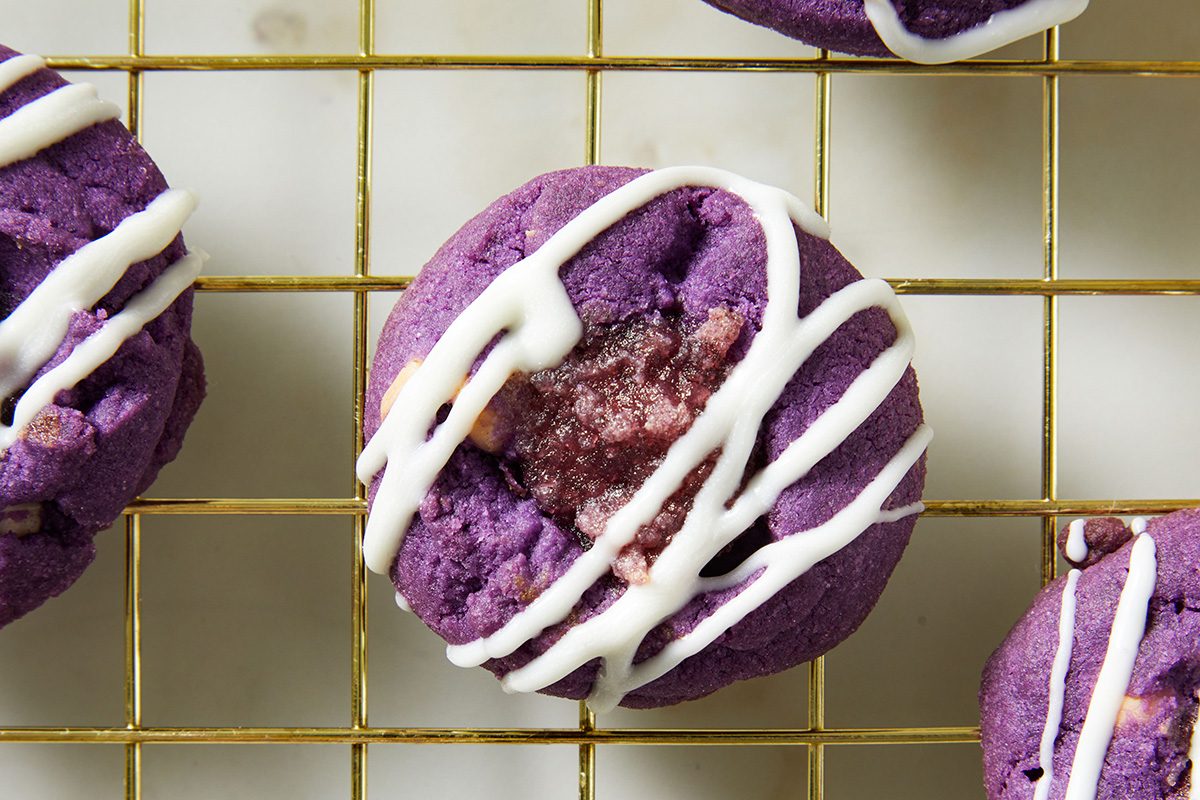 A vibrant purple cookie with a gooey center and drizzled with white icing, resting on a gold wire cooling rack.