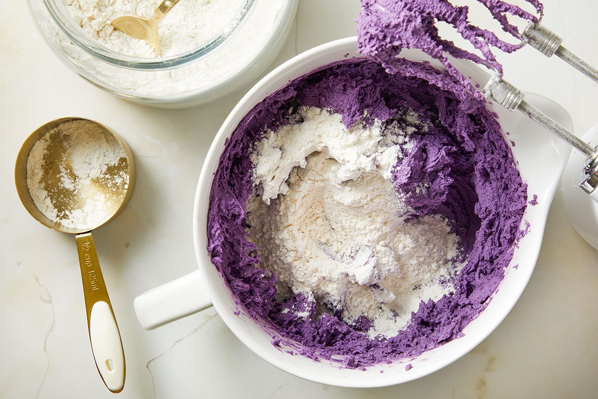 A white mixing bowl contains vibrant purple batter with flour on top. An electric hand mixer with batter on the beaters rests on the bowl’s edge. Nearby are a gold measuring cup and a glass jar of flour.