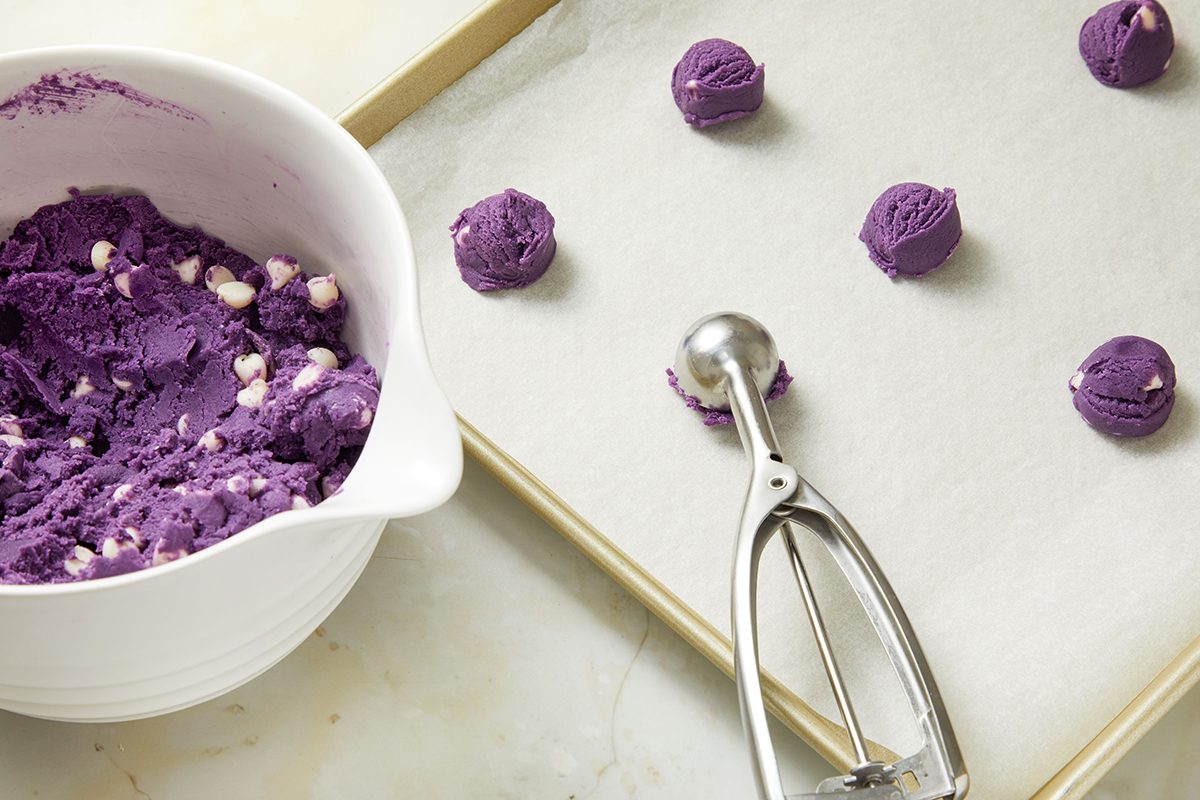 A baking sheet with scoops of purple cookie dough, some placed on parchment paper, sits next to a bowl of the same dough with white chocolate chips and a metal scoop.