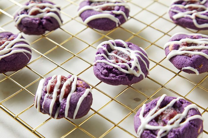 Eight vibrant purple cookies with white icing drizzle are cooling on a gold wire rack, placed on a white surface.