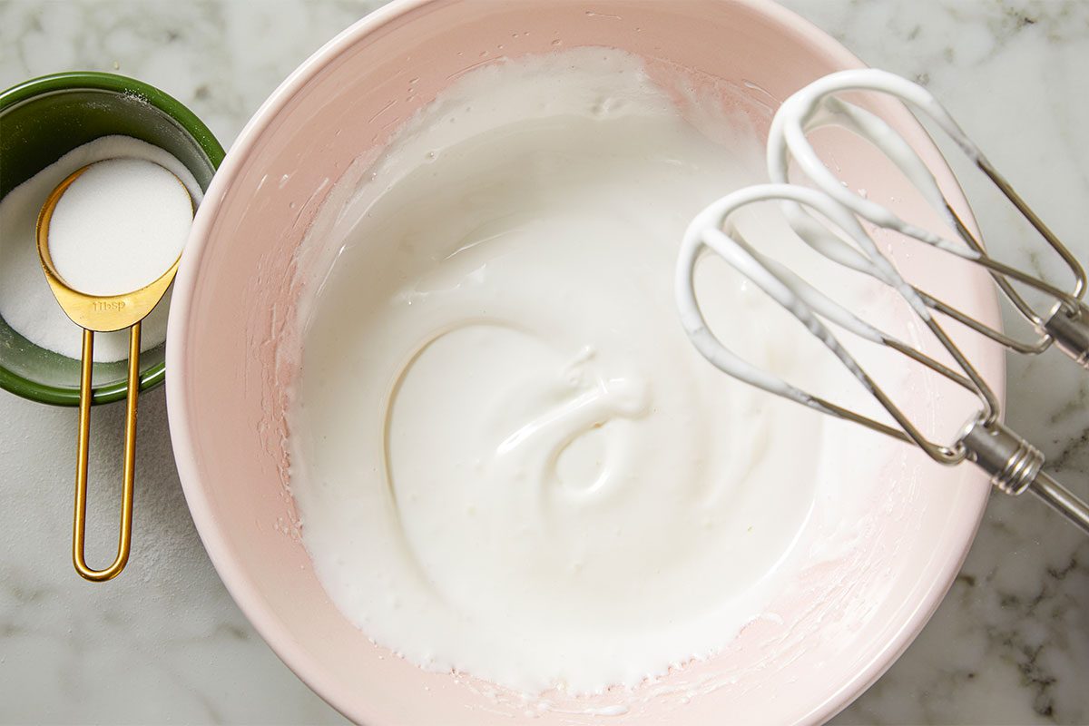Overhead shot of a pink bowl holds shiny whipped meringue on a marble surface next to a hand mixer with beaters and a measuring cup of sugar
