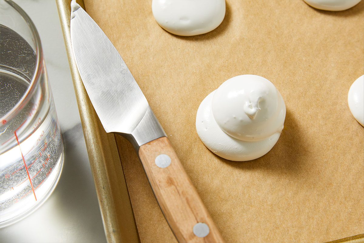 Overhead shot of a baking tray with parchment paper is topped with white meringue dollops a wooden-handled knife rests nearby and next to it sits a glass measuring cup filled with water