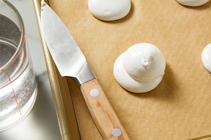 Overhead shot of a baking tray with parchment paper is topped with white meringue dollops a wooden-handled knife rests nearby and next to it sits a glass measuring cup filled with water