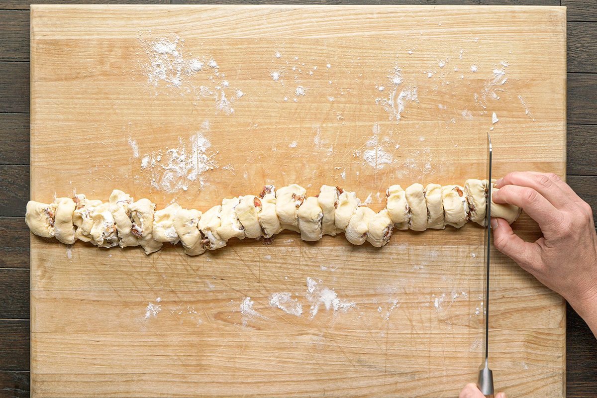 Close-up shot; a hand slices a long; twisted cinnamon-filled dough roll on a floured wood surface