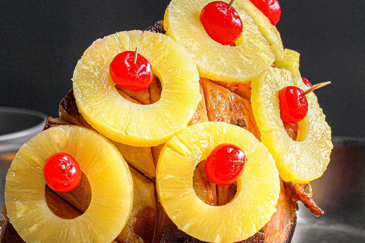 A baked ham garnished with circular pineapple rings and bright red maraschino cherries, arranged decoratively on the surface.