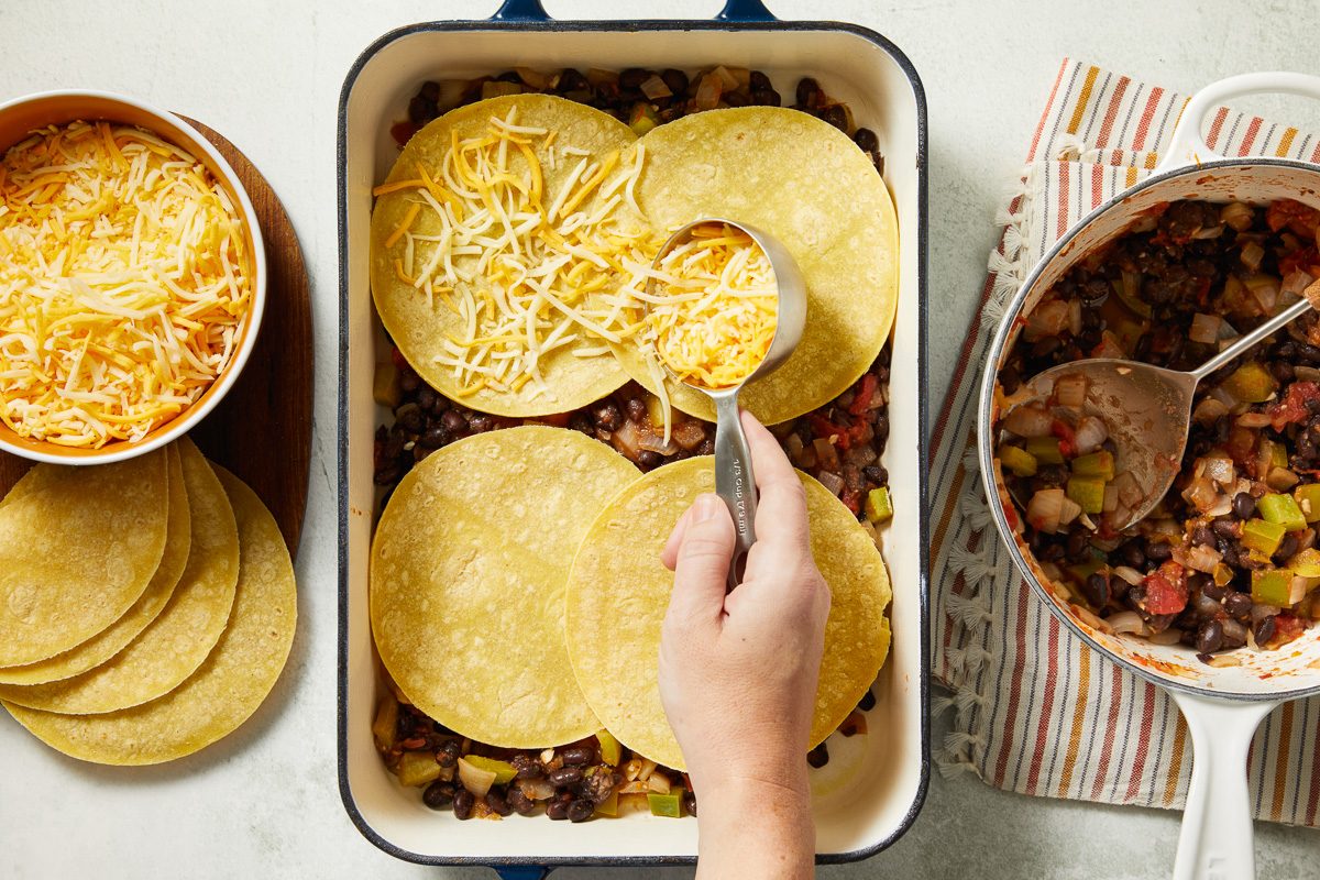 cheese being sprinkled on tortillas layered on top of veggie mixture in a baking dish