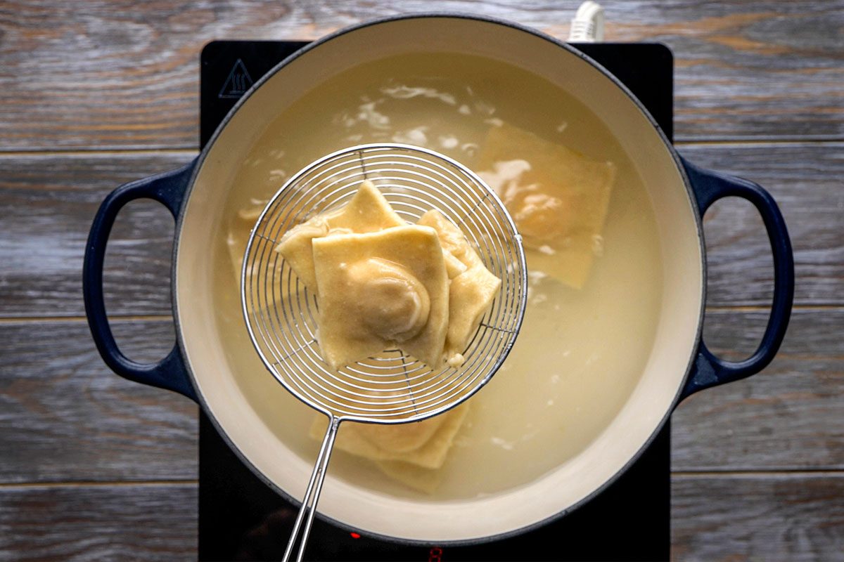 Overhead shot of a metal strainer lifts ravioli from a pot of boiling water on a stovetop and the wooden surface is in the background nearby
