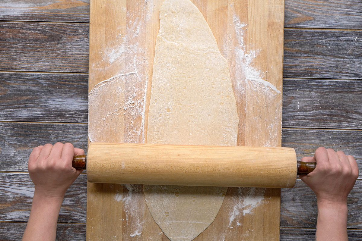Overhead shot of a hands roll out dough with a pin on a floured wooden surface and cutting board the scene shows baking preparations in progress