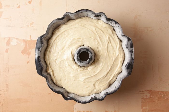 Overhead shot of a bundt cake pan holds smooth unbaked cake batter on a light tan textured surface and the pan is fluted and lightly coated with flour