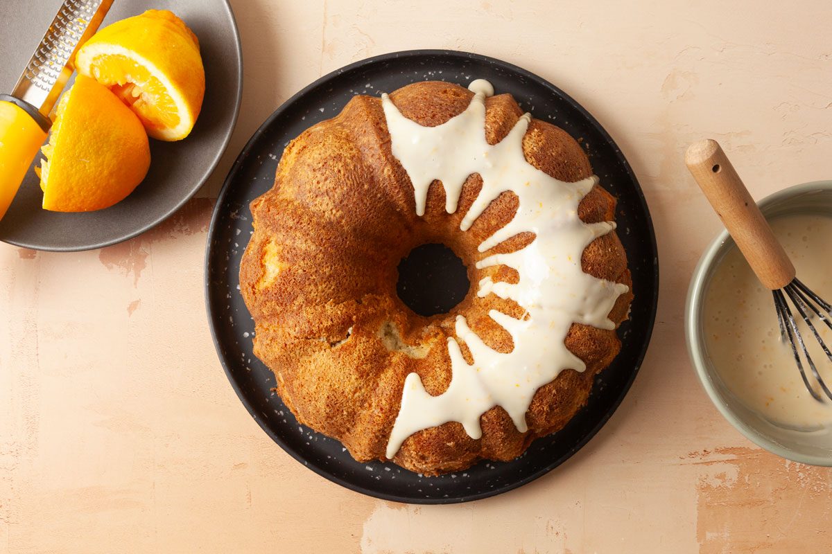 Overhead shot of Cardamom Pound Cake with white icing drizzled on top rests on a dark plate beside it are glaze with a whisk and orange slices in the background is a light tan surface