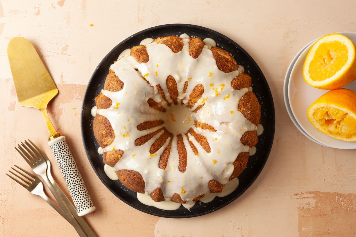 Overhead shot of Cardamom Pound Cake with orange zest on top is placed on a black plate with a gold cake server forks and halved oranges nearby the scene rests on a beige surface