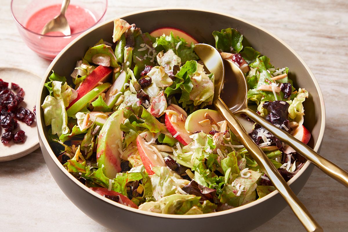 3/4 angle view shot of Merry Berry Salad with crisp greens, sliced apples, dried cranberries, and shredded cheese in a beige bowl with gold utensils; A bowl of pink berry dressing and a small plate of cranberries sit alongside on a light marble surface;