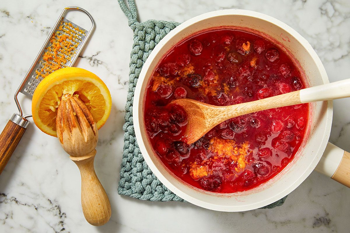 Overhead shot of cooked cranberries in a saucepan with zest and juice from a freshly squeezed lemon, wooden spoon resting in the thickening mixture