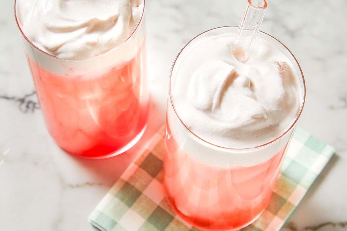 Angled overhead shot of two tall glasses of cranberry cream ginger ale, showcasing the ombré pink drink and thick whipped topping.