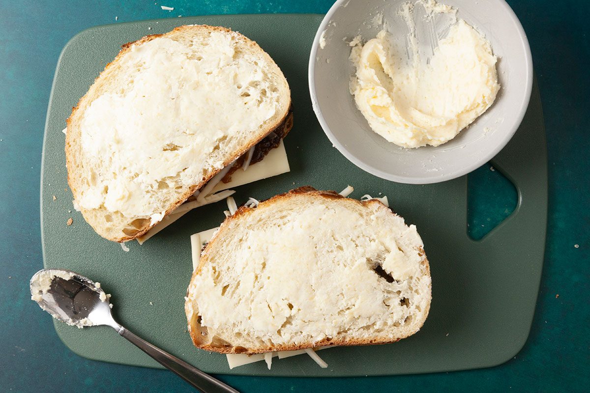 Overhead shot of two slices of bread spread generously with a creamy butter mixture on a cutting board; A small bowl of the spread and a spoon rest nearby on a teal surface, as the sandwiches are being assembled;