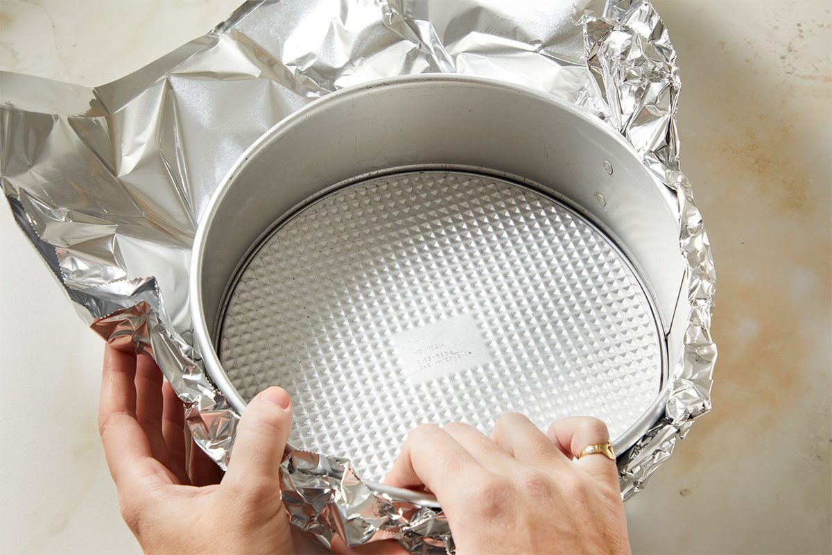 Overhead shot of a springform pan wrapped in foil, hands pressing the foil tightly around the base to prepare it for a water bath.