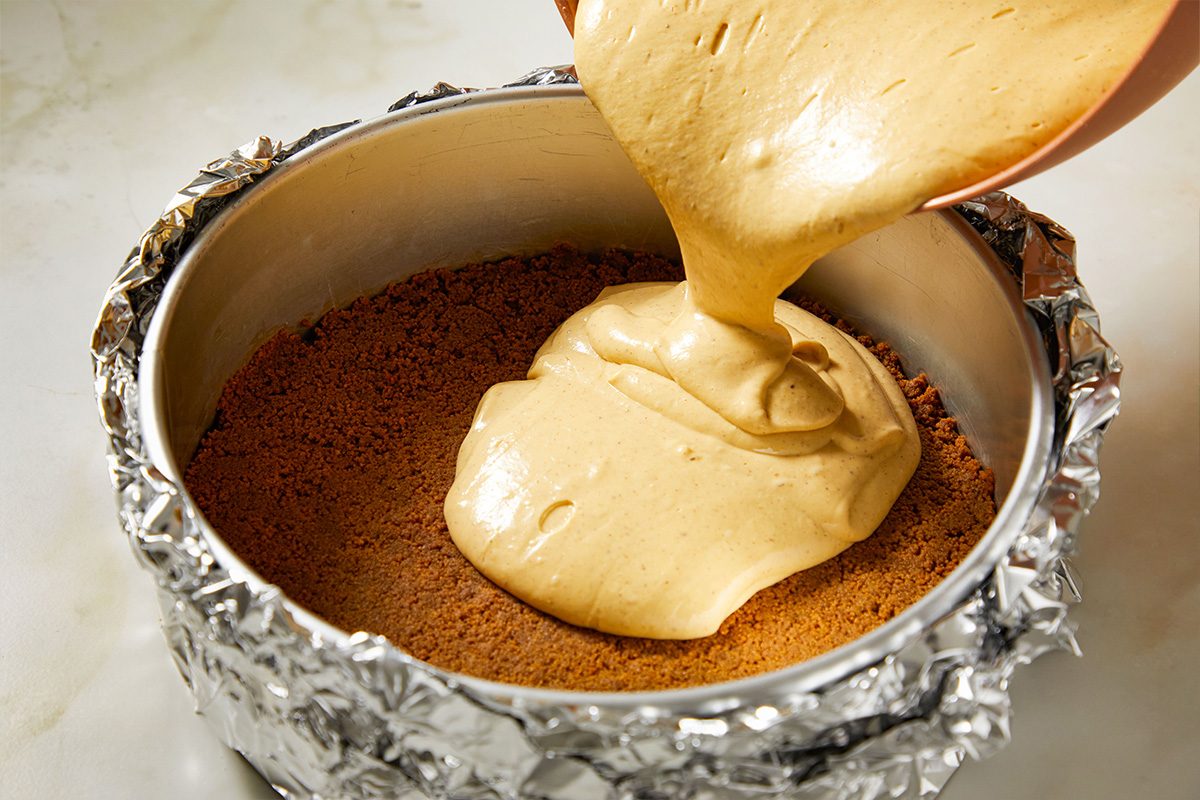 Overhead shot of silky gingerbread cheesecake batter being poured over the pressed crust inside the foil-wrapped springform pan.