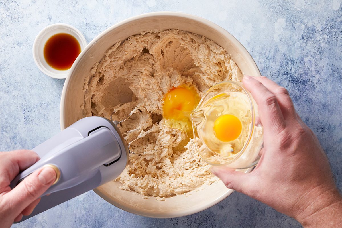 Overhead shot of a person adds eggs into dough in a mixing bowl using an electric hand mixer and there is a small bowl of vanilla extract nearby on a blue countertop