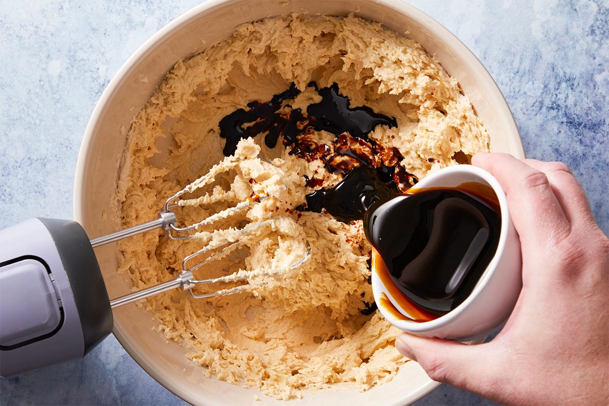 Overhead shot of a hand pours dark molasses into a mixing bowl with creamy dough and an electric mixer sits inside the bowl on a light blue surface