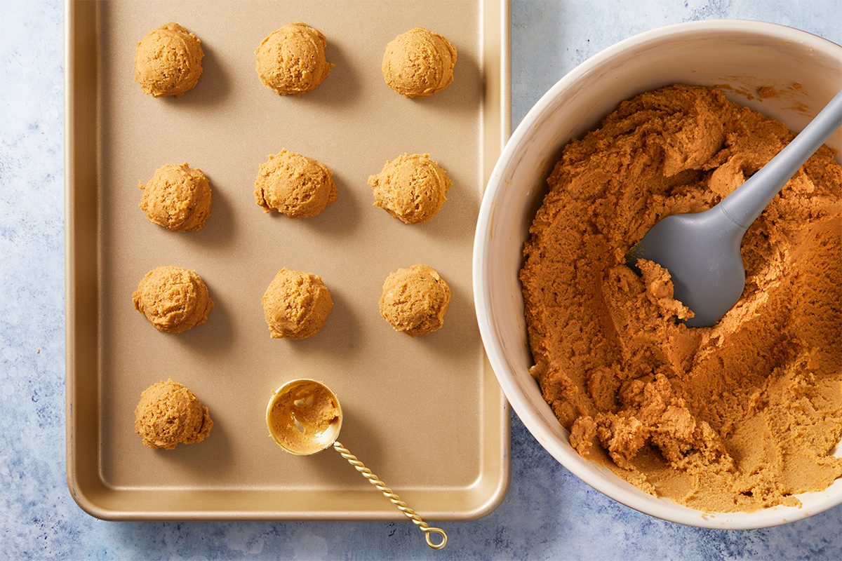 Overhead shot of a baking sheet has twelve scoops of cookie dough and it rests near a bowl filled with more dough with a gray spatula and gold scoop both are placed on a light blue surface