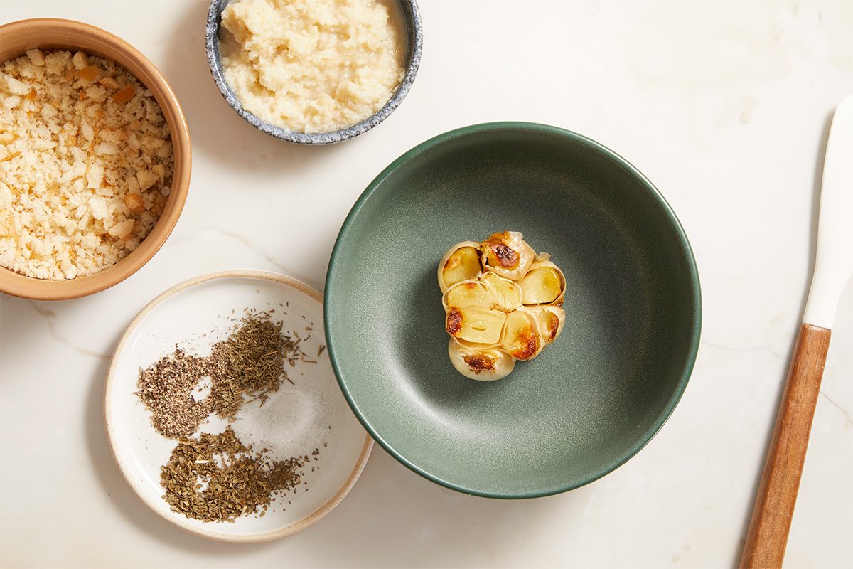 A green bowl with a roasted garlic bulb, surrounded by dishes containing breadcrumbs, shredded cheese, and dried herbs, all on a light-colored surface with a knife nearby.